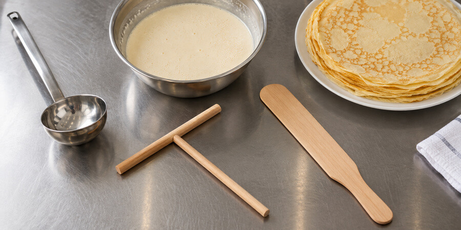 Wooden crepe spreader and spatula beside a ladle, bowl of batter, and stack of finished crepes.