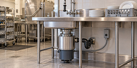 Commercial food waste disposer mounted beneath a stainless steel sink in a restaurant dishroom.