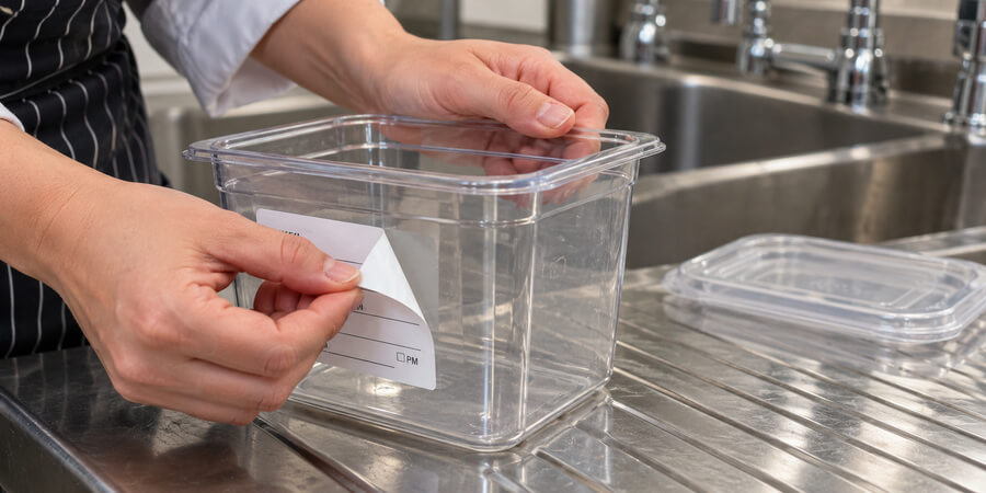 Hands peeling a removable food rotation label off a clear container at the sink
