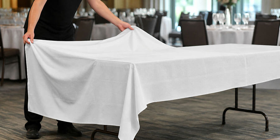 Staff spreading a white tablecloth over a rectangular folding table in a banquet room