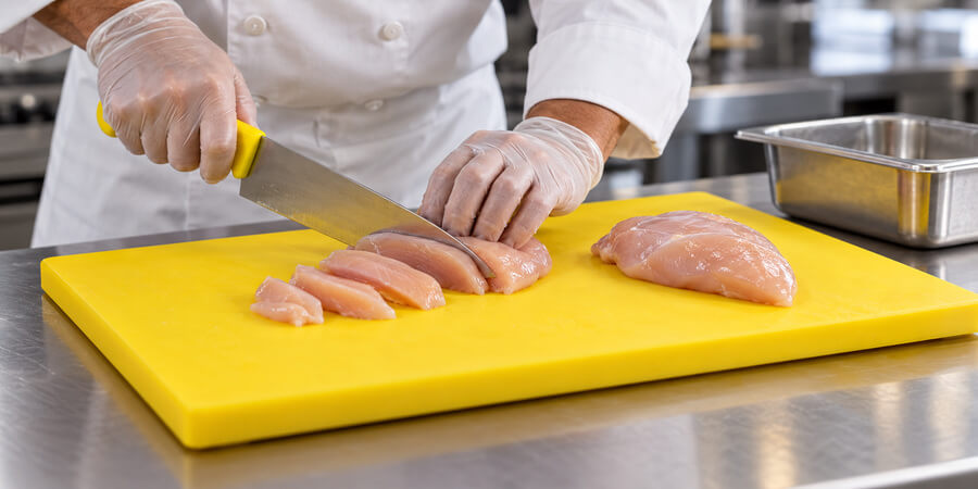 Cook in gloves slicing raw chicken breast on a yellow cutting board